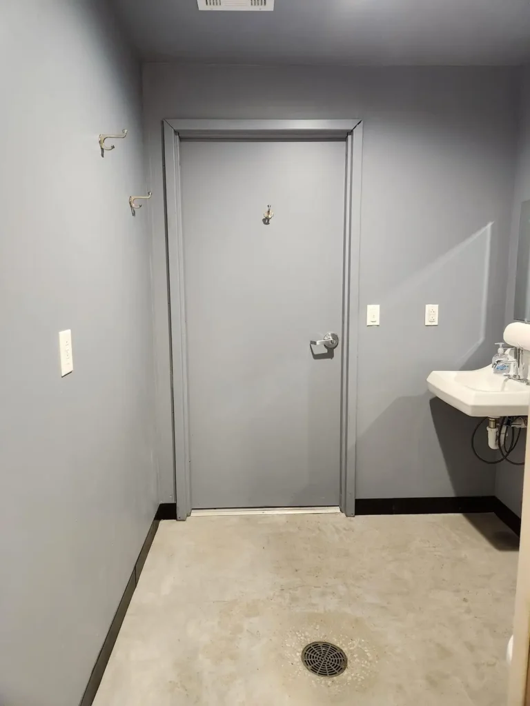 Plain gray restroom or shower room interior with metal door, wall hooks, small white sink, and concrete floor drain.