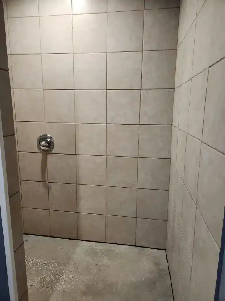 Empty beige tiled shower stall with a single chrome mixer control valve and a bare concrete floor.