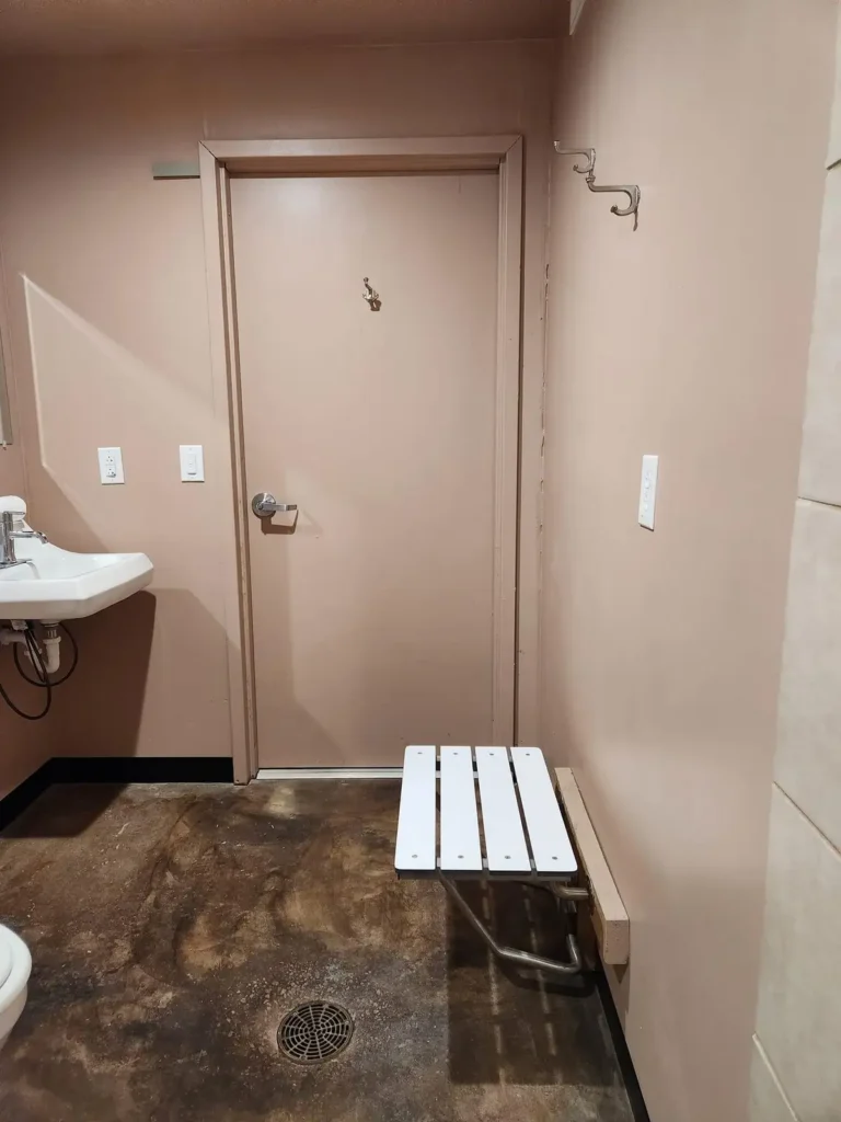 Accessible shower room interior with fold-down white slat bench, wall-mounted sink, floor drain, and metal door with