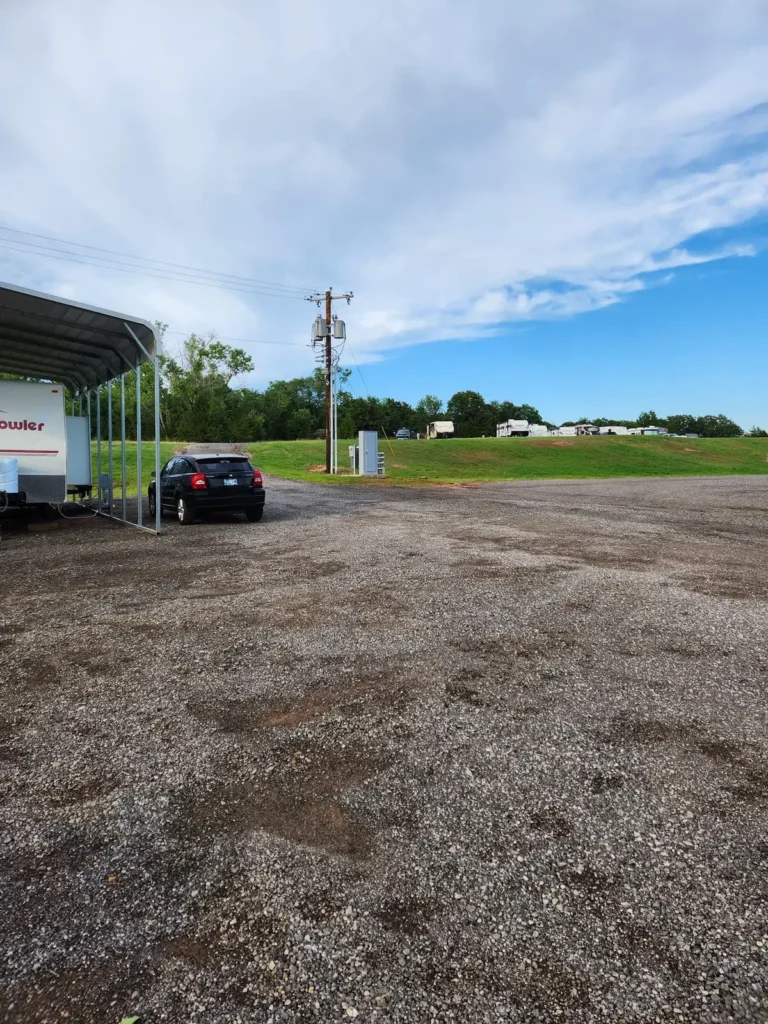 Wide gravel lot with a black hatchback parked under a metal carport beside a Fleetwood travel trailer, RVs on the